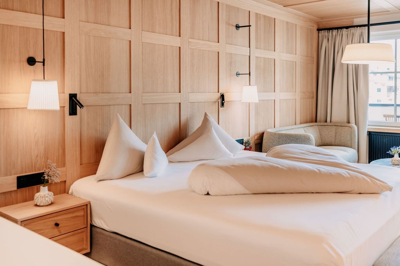 Natursuite „Lebensquell“ mit Holzverkleidung an der Wand, Schlafzimmer und angrenzendem Wohnbereich mit Sofa – harmonisches, naturinspiriertes Interior-Design.