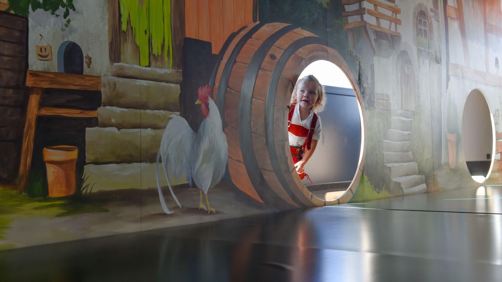 Kind spielt im Baby- und Kleinkinderbereich „Muckasackal“ des ULRICHSHOF und schaut lachend aus einem großen Holzfass. Die Wandmalerei mit Bauernhof-Szene und Hahn sorgt für eine bunte, kindgerechte Atmosphäre.