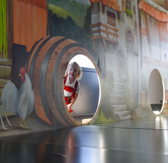 Kind spielt im Baby- und Kleinkinderbereich „Muckasackal“ des ULRICHSHOF und schaut lachend aus einem großen Holzfass. Die Wandmalerei mit Bauernhof-Szene und Hahn sorgt für eine bunte, kindgerechte Atmosphäre.