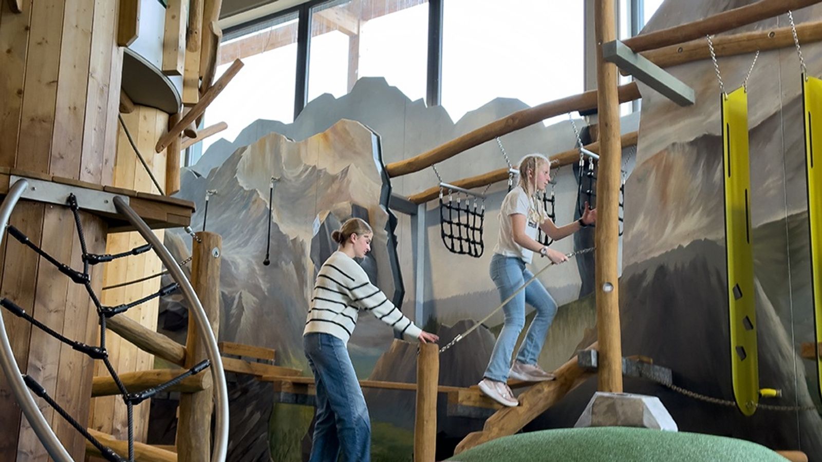 Zwei Kinder klettern in der Spiel-KISTE des ULRICHSHOF über ein abenteuerliches Klettergerüst aus Holz mit Seilen und Brücken, während im Hintergrund eine Berglandschaft auf der Wand gemalt ist.