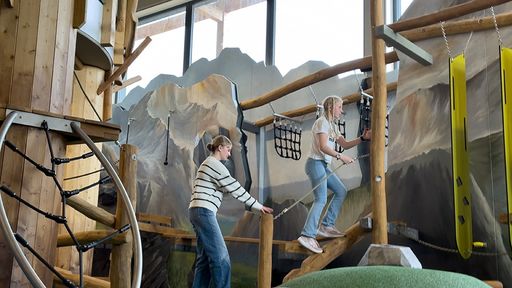 Zwei Kinder klettern in der Spiel-KISTE des ULRICHSHOF über ein abenteuerliches Klettergerüst aus Holz mit Seilen und Brücken, während im Hintergrund eine Berglandschaft auf der Wand gemalt ist.