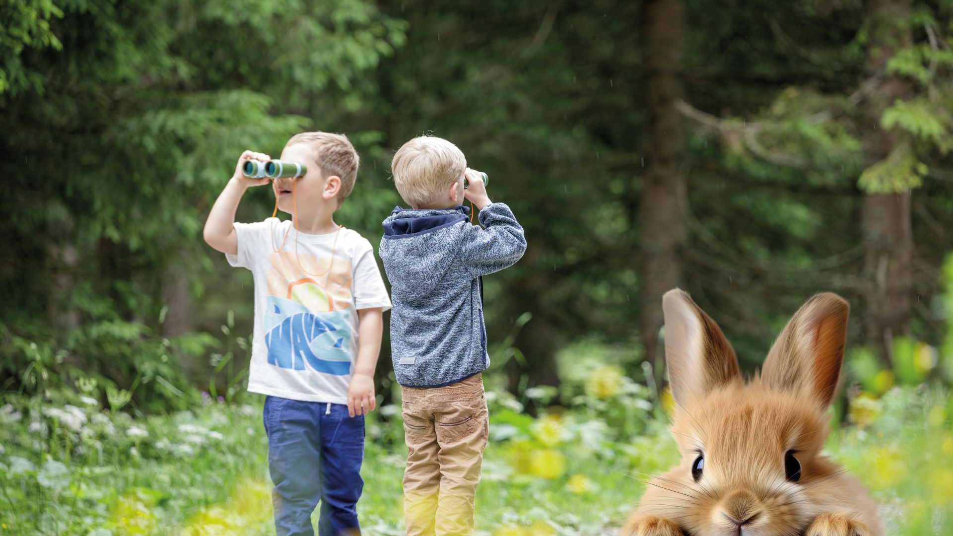 Two children stand on a forest path, looking into the distance with binoculars, while a curious Easter bunny peeks out from the grass in the foreground.