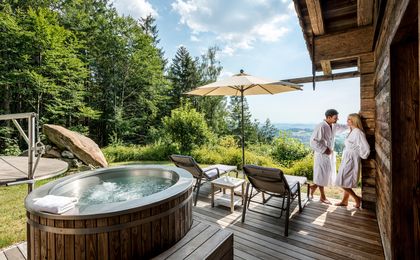 Pärchen steht im Luxus-Chalet auf der eigenen Sonnenterrasse neben Whirlpool und traumhaften Ausblick über den Bayerischen Wald.