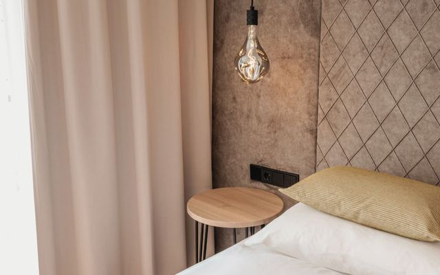 Ein stilvolles Schlafzimmer-Detail im Hotel die HOCHKÖNIGIN in Maria Alm: edles Kopfteil mit Steppmuster, sanfte Stoffe, natürliche Farben und eine Designerleuchte, die Gemütlichkeit mit modernem Stil verbindet.