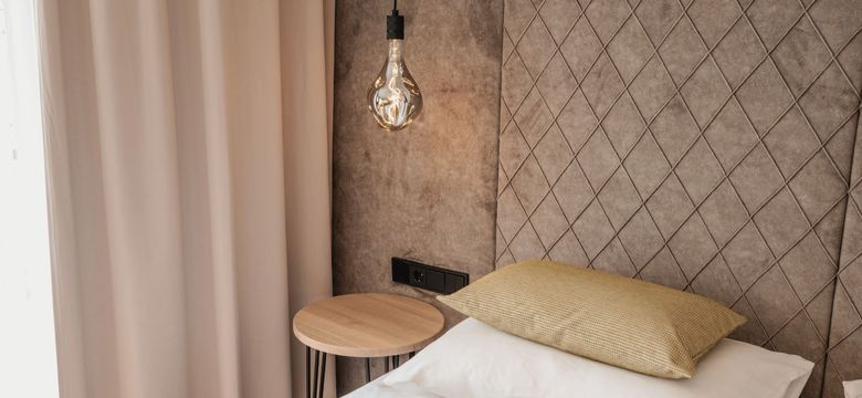 Ein stilvolles Schlafzimmer-Detail im Hotel die HOCHKÖNIGIN in Maria Alm: edles Kopfteil mit Steppmuster, sanfte Stoffe, natürliche Farben und eine Designerleuchte, die Gemütlichkeit mit modernem Stil verbindet.