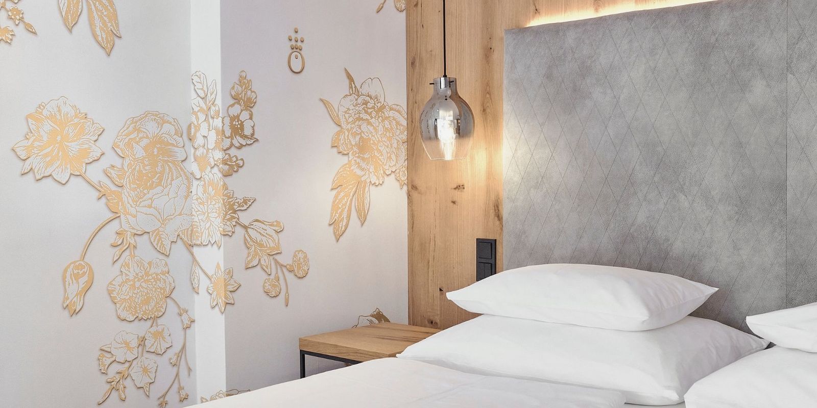 Modernes und edles Zimmerdetail im Hotel die HOCHKÖNIGIN in Maria Alm: kunstvolles Wanddesign in Gold, hochwertige Stoff-Polsterwand, warme Holzdetails und stilvolle Hängeleuchte. Perfekt für Gäste, die modernes Design und besondere Wohlfühlmomente lieben.