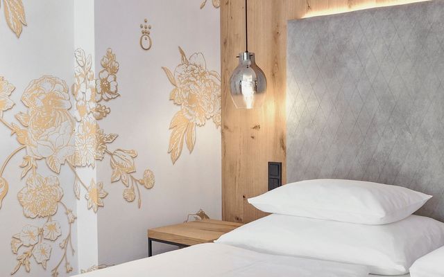 Modernes und edles Zimmerdetail im Hotel die HOCHKÖNIGIN in Maria Alm: kunstvolles Wanddesign in Gold, hochwertige Stoff-Polsterwand, warme Holzdetails und stilvolle Hängeleuchte. Perfekt für Gäste, die modernes Design und besondere Wohlfühlmomente lieben.