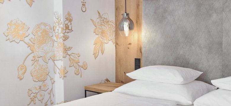 Modernes und edles Zimmerdetail im Hotel die HOCHKÖNIGIN in Maria Alm: kunstvolles Wanddesign in Gold, hochwertige Stoff-Polsterwand, warme Holzdetails und stilvolle Hängeleuchte. Perfekt für Gäste, die modernes Design und besondere Wohlfühlmomente lieben.
